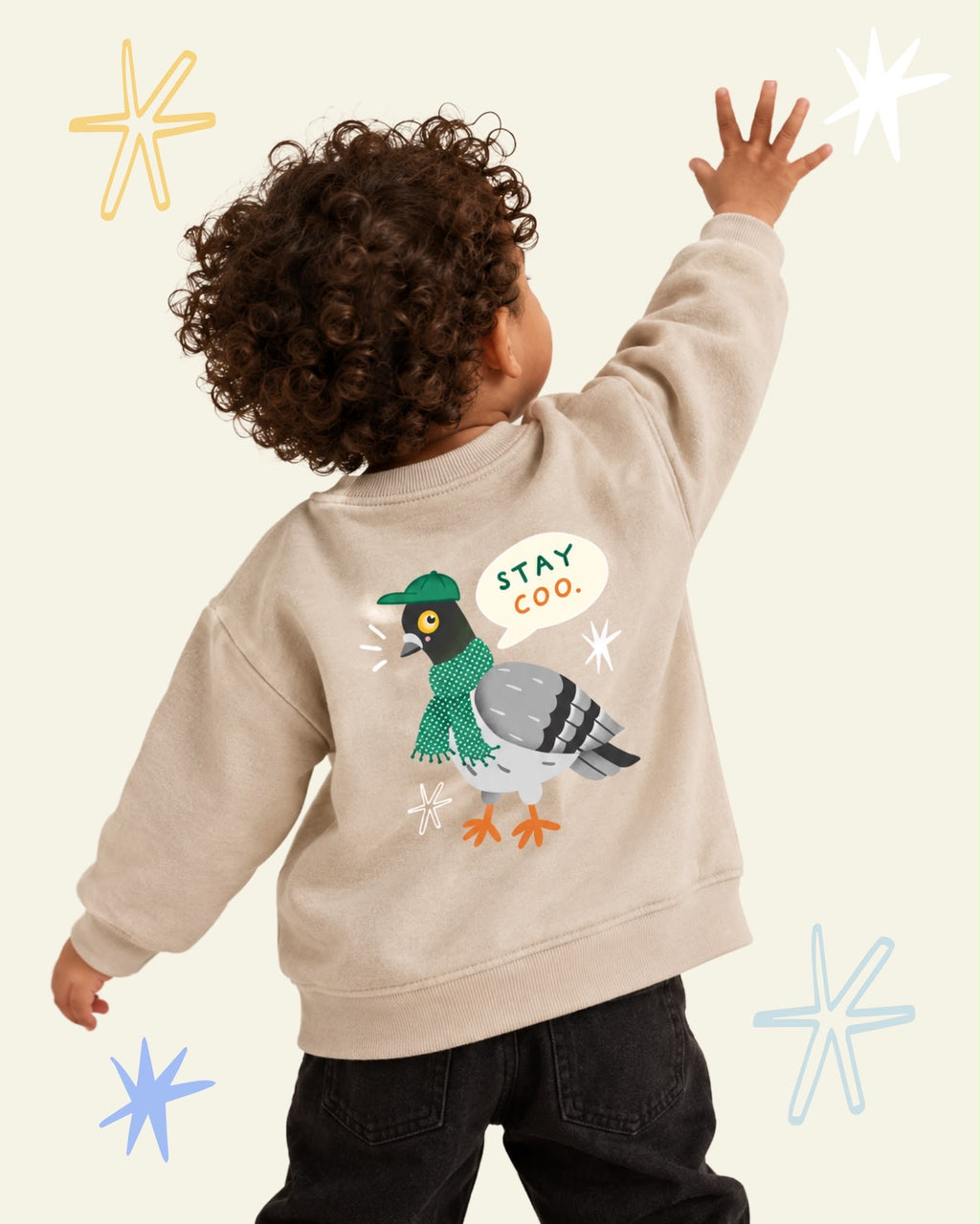 Child wearing a beige Stay Coo. sweatshirt with a pigeon in a green cap and scarf printed on the back.