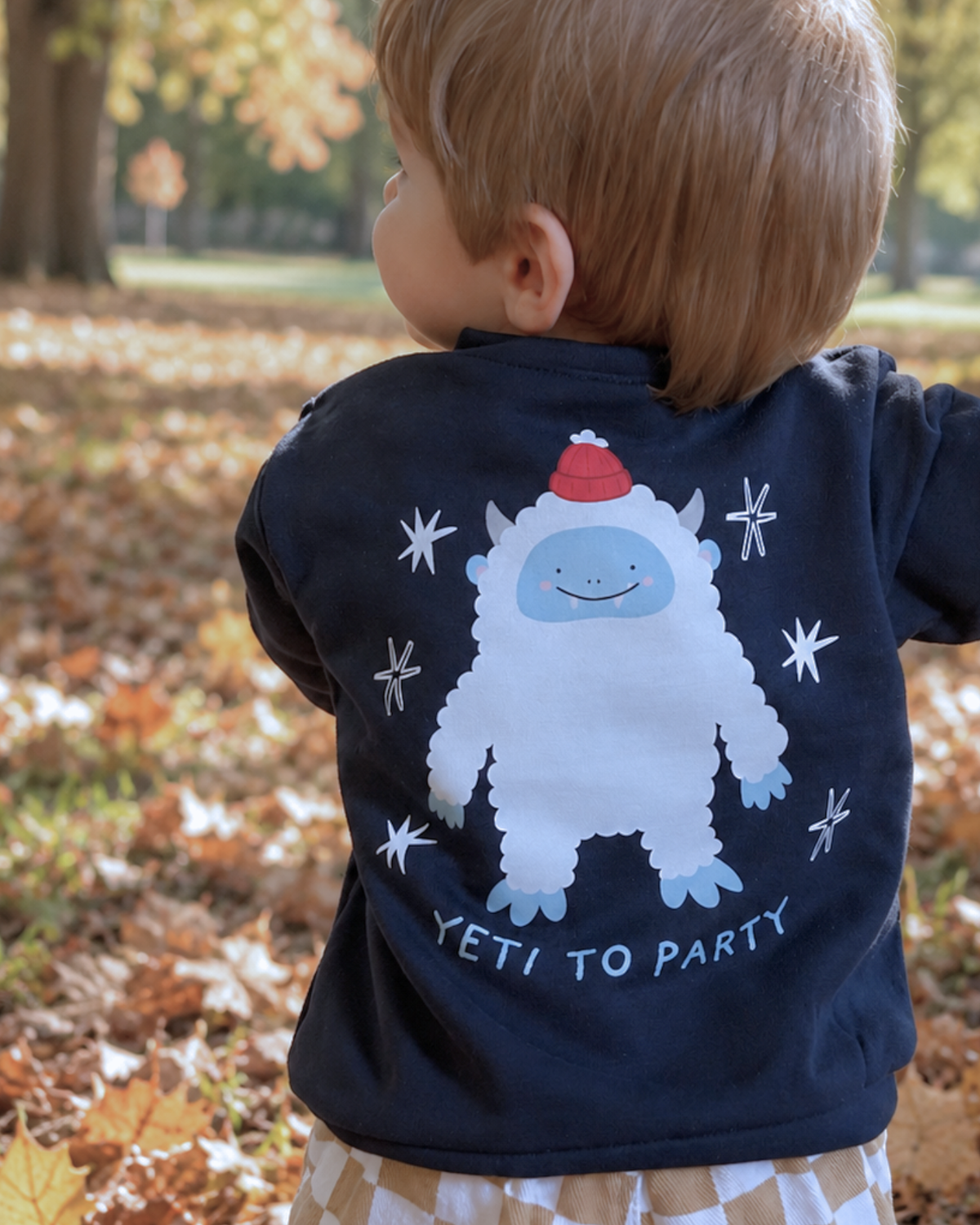 Child among autumn leaves wearing a navy Yeti to Party sweatshirt, showing the white yeti back print.