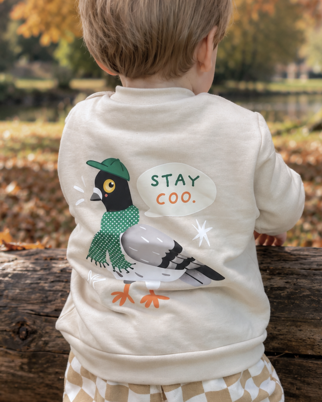 Child outdoors wearing a light-coloured Stay Coo. sweatshirt, showing the pigeon back print with green cap and scarf.